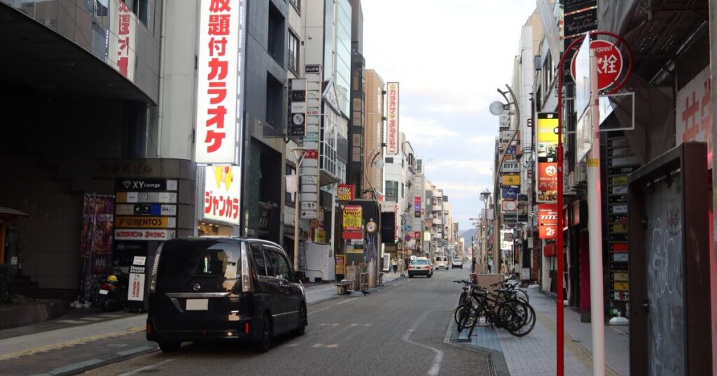 看板が並ぶ繁華街の通りと走行する車のある都市風景